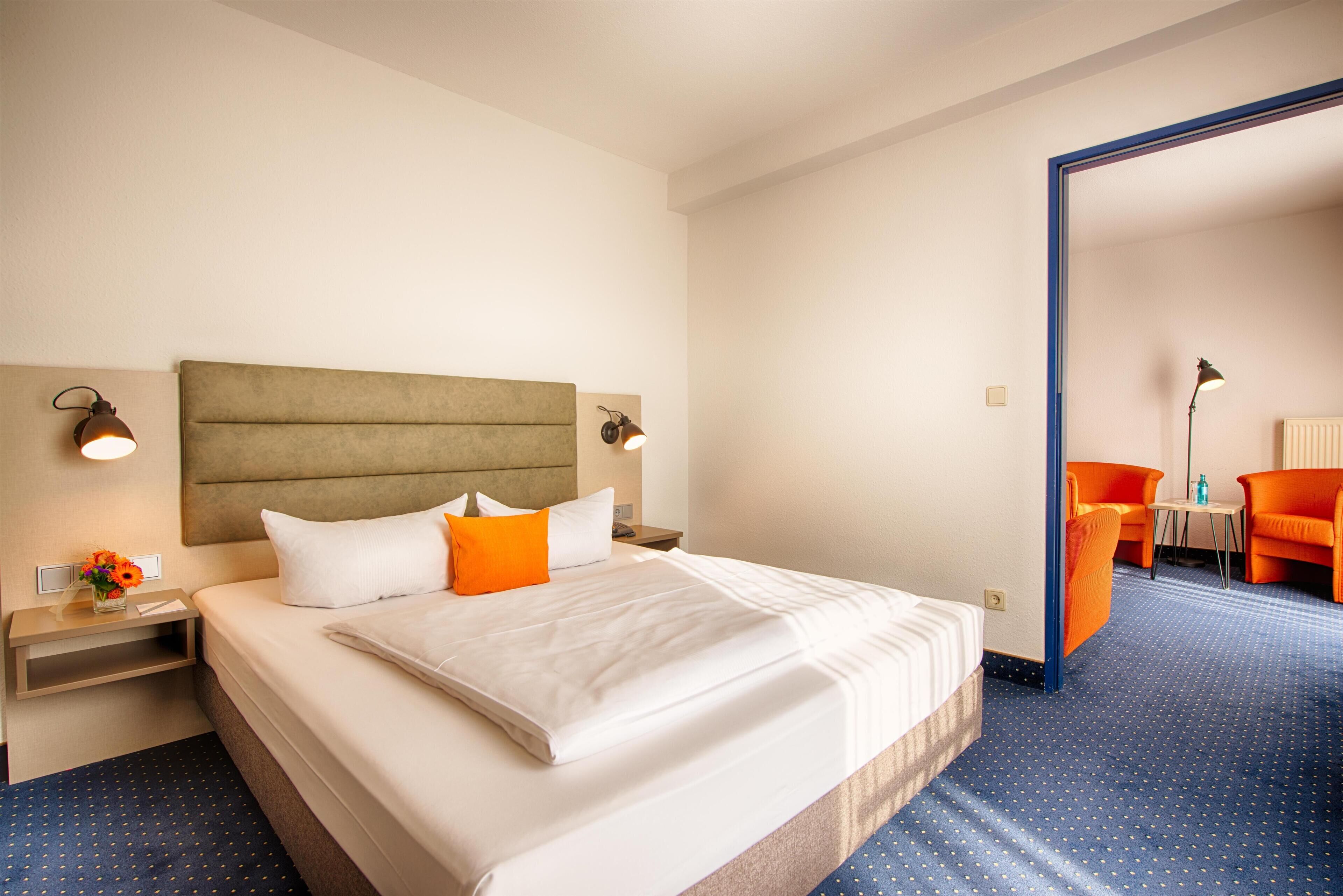 Habitación de hotel con una cama grande, dos mesitas de noche, un escritorio y una silla naranja, sobre una alfombra azul.