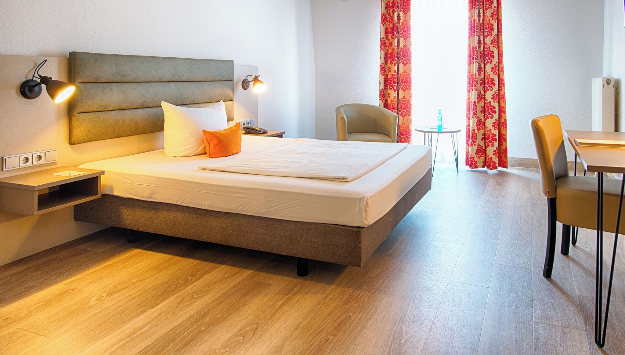 A hotel room with a bed, bedside tables, a chair, a small table, and a desk.