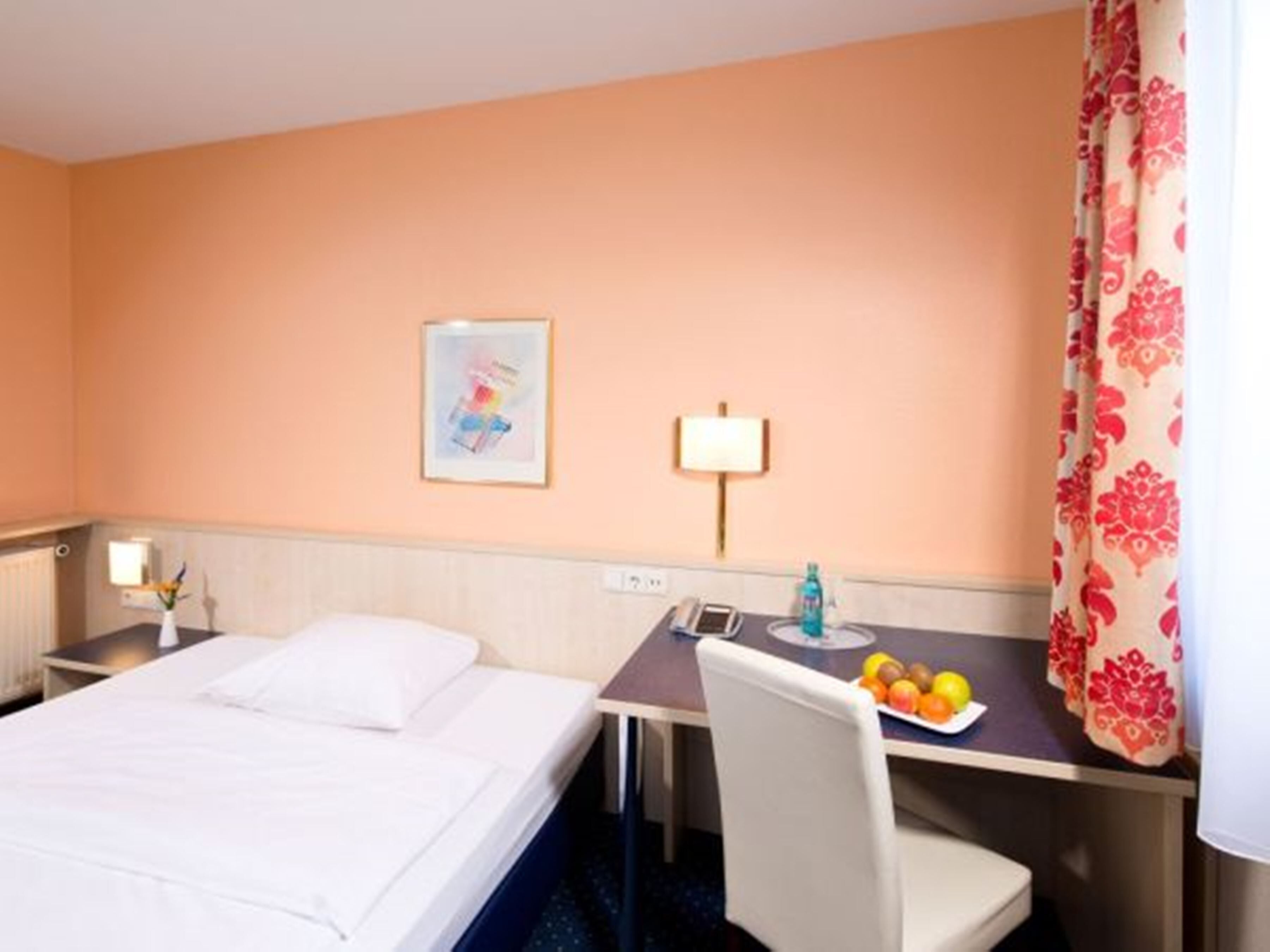 A hotel room with a bed, a desk with a chair, a lamp, a framed picture on the wall, and a fruit plate on the desk.