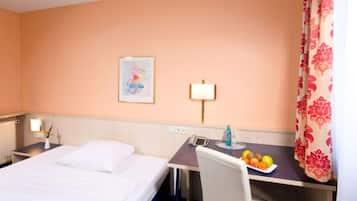 A hotel room with a bed, a desk with a chair, a lamp, a framed picture on the wall, and a fruit plate on the desk.