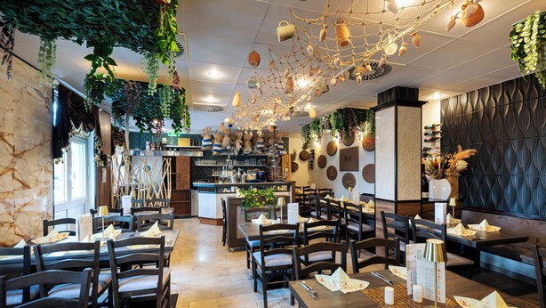 Un restaurant avec des tables et des chaises en bois, un espace cuisine et des plantes suspendues.