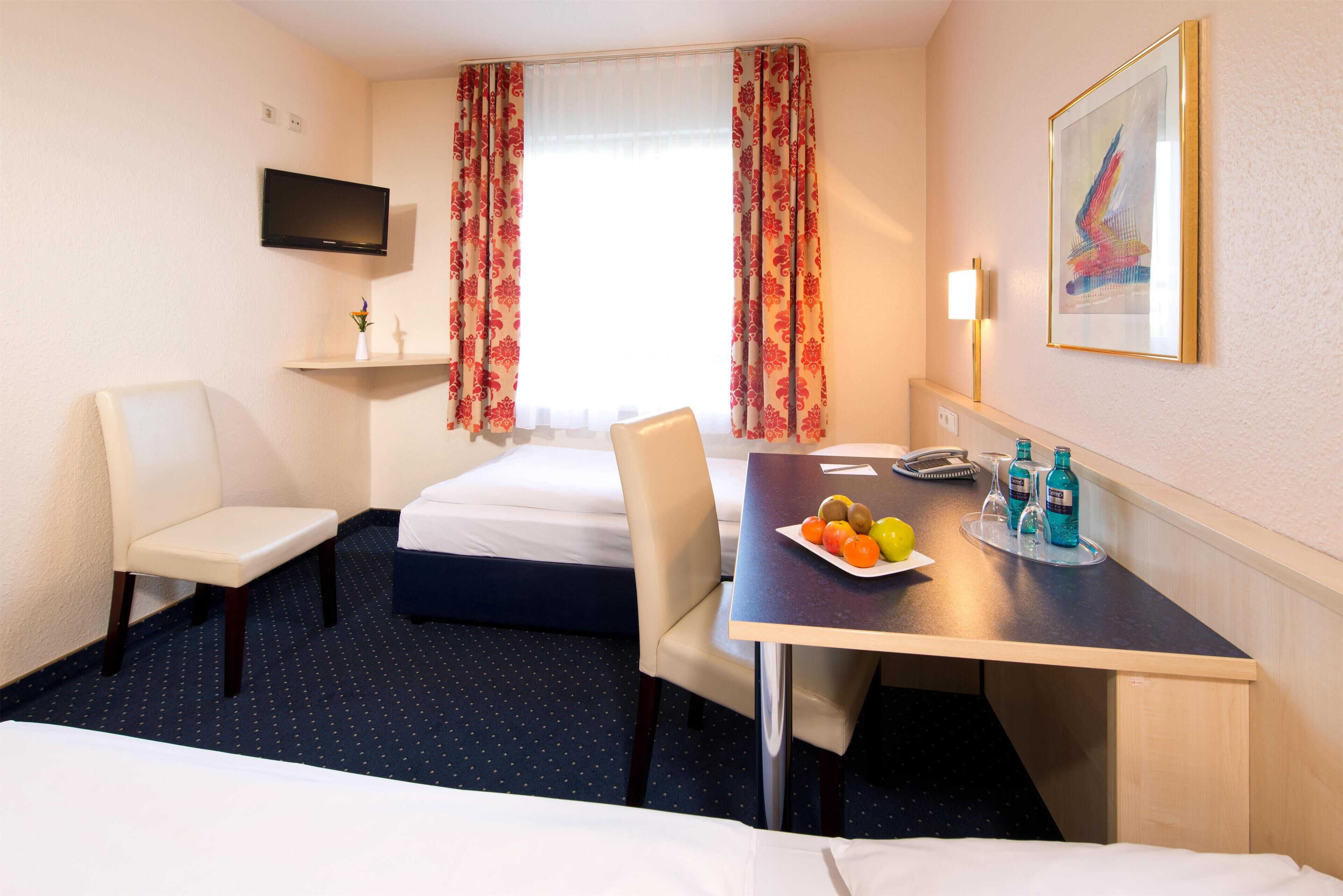 A hotel room with a bed, two chairs, a desk with a plate of fruit and bottles, a TV, and a painting on the wall.