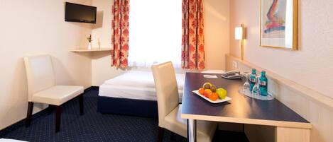 A hotel room with a bed, two chairs, a desk with a plate of fruit and bottles, a TV, and a painting on the wall.