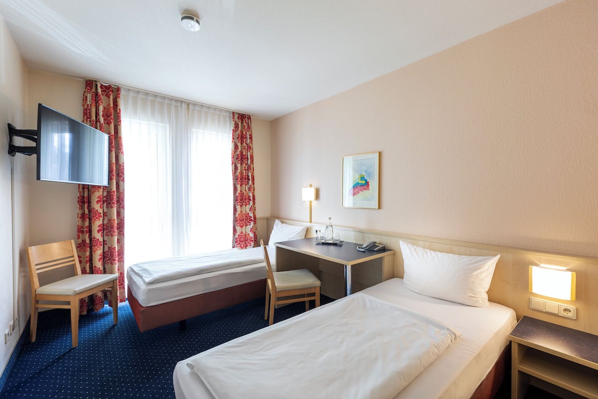 Quarto de hotel com duas camas, uma escrivaninha, uma cadeira, uma televisão e uma janela com cortinas.