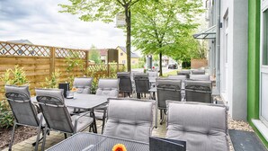 Un espace extérieur aménagé en terrasse, avec plusieurs tables et chaises, entouré d’une clôture en bois et de verdure.