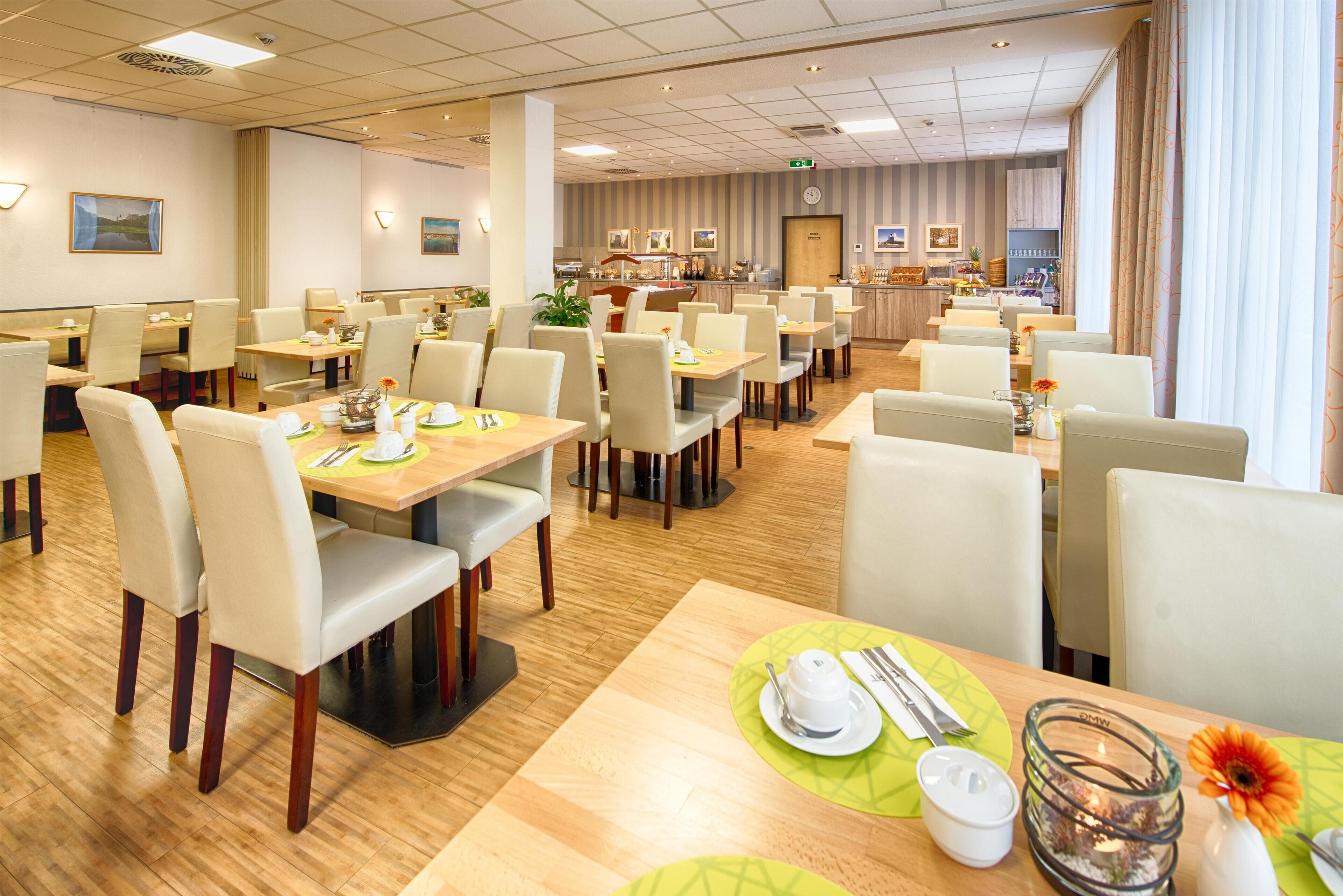 A spacious hotel dining area with multiple tables and chairs, a kitchen in the background, and a view of the city through large windows.