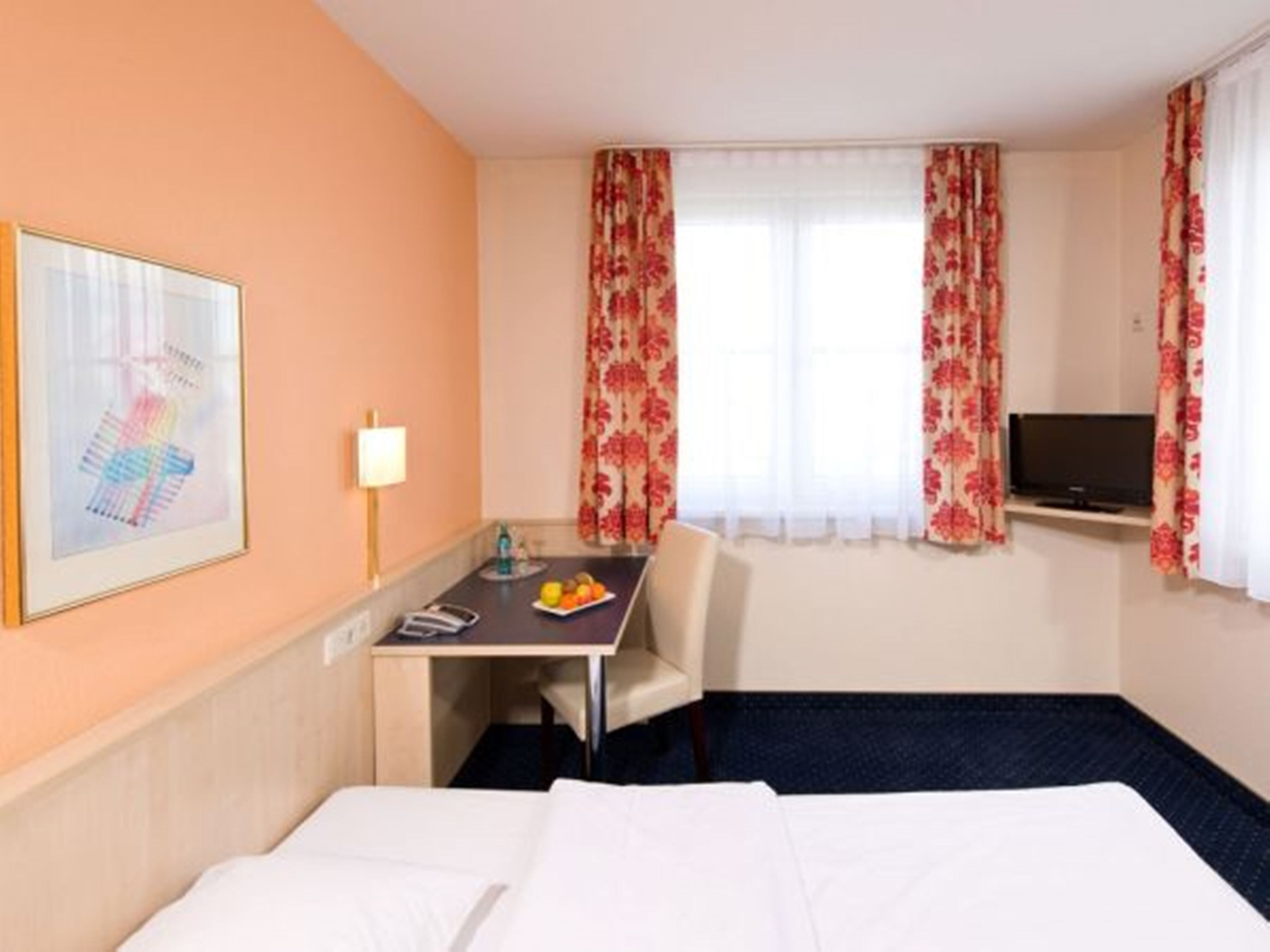 A hotel room with a bed, a desk with a chair, a television, and a window with floral curtains.