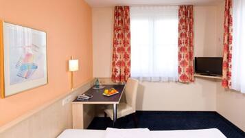 A hotel room with a bed, a desk with a chair, a television, and a window with floral curtains.