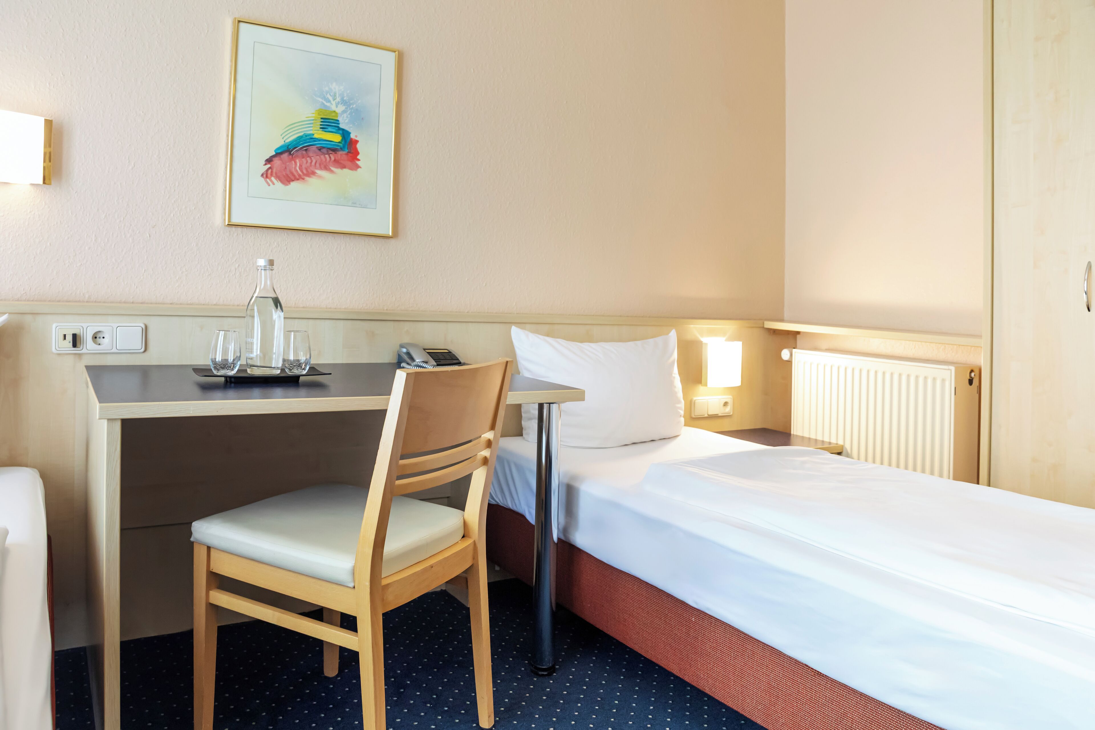 Une chambre d’hôtel avec un lit, un bureau, une chaise, une lampe, un tableau et un radiateur.