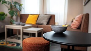 Un salon avec une table ronde noire, un pouf marron, un bol gris rempli d’en-cas, une table d’appoint blanche et un canapé marron avec des coussins.