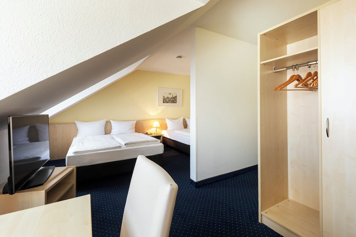 Quarto de hotel com cama, mesa de cabeceira, abajur e guarda-roupa de madeira.