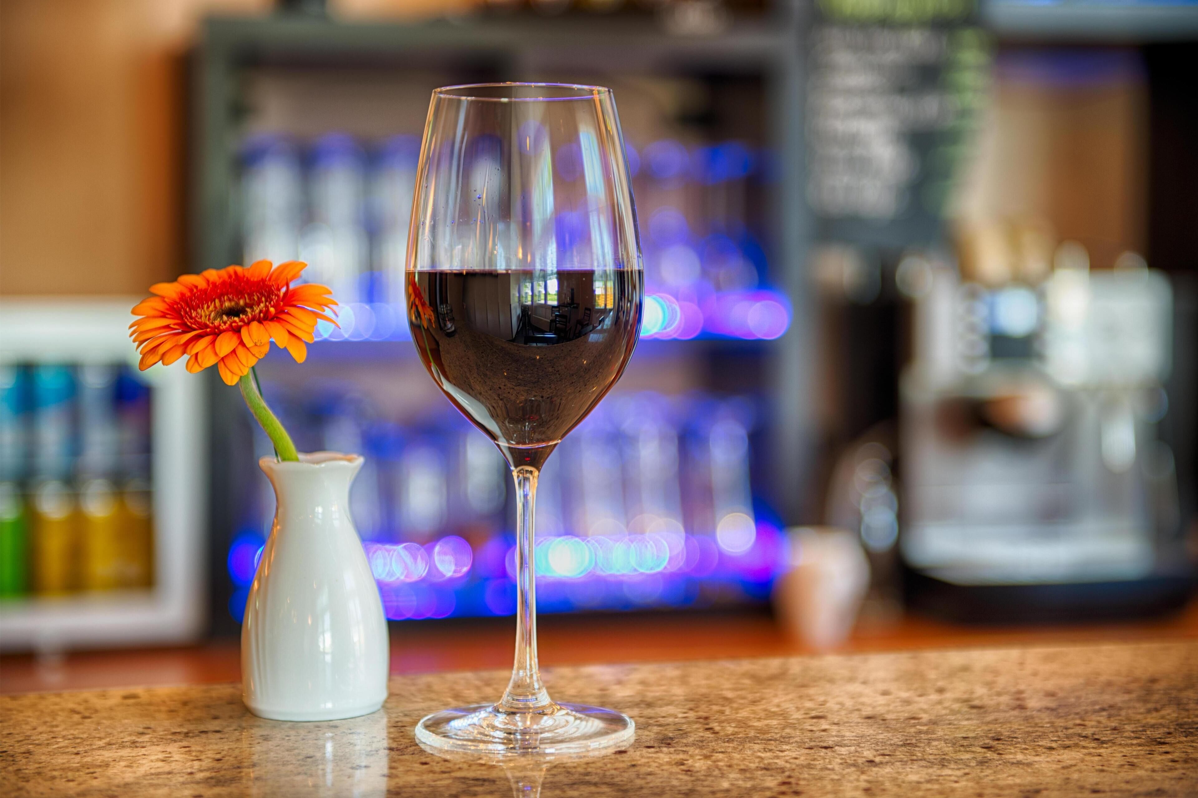 A glass of red wine and a vase with a single orange flower on a counter.
