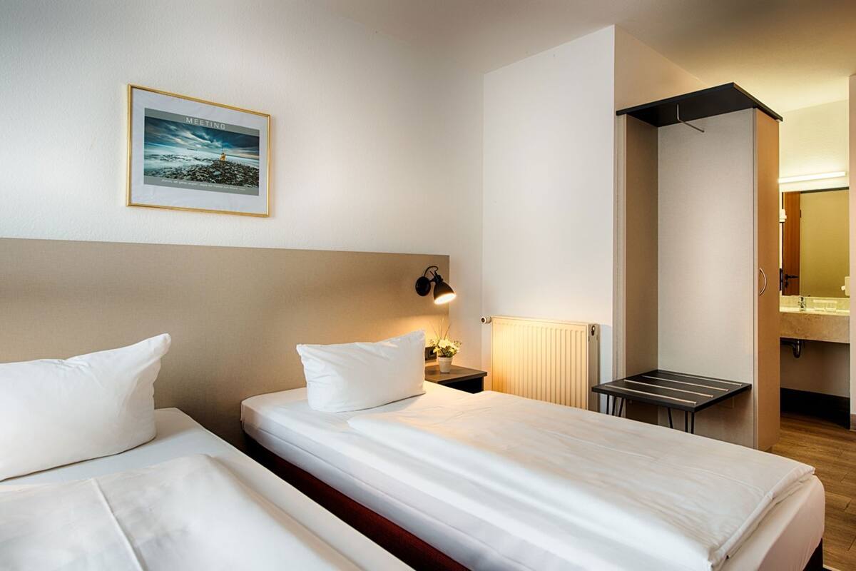 A hotel room with two beds, a bedside table with a lamp, a radiator, and a framed picture on the wall.