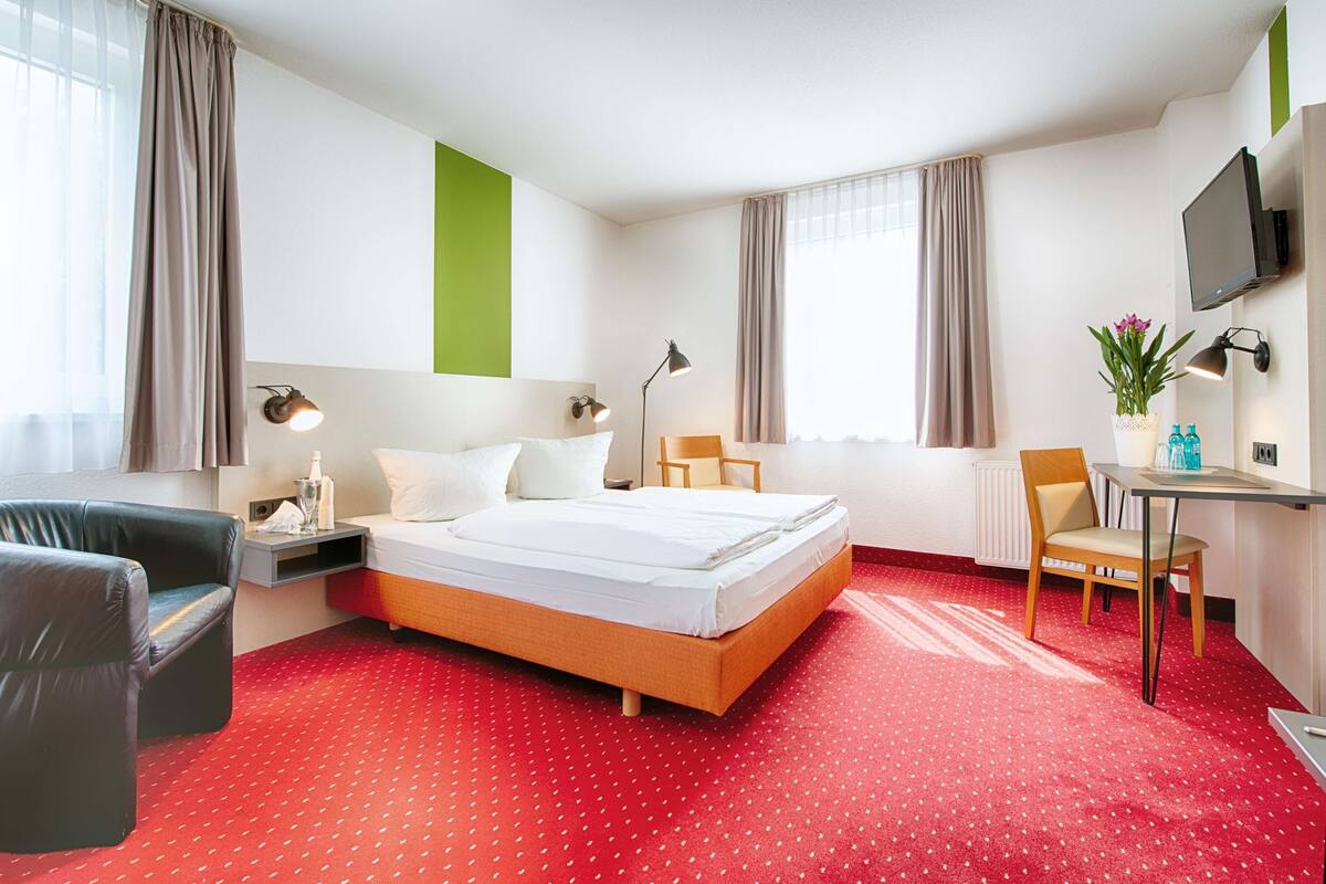 A hotel room with a bed, a desk, a chair, a TV, and a window with curtains.