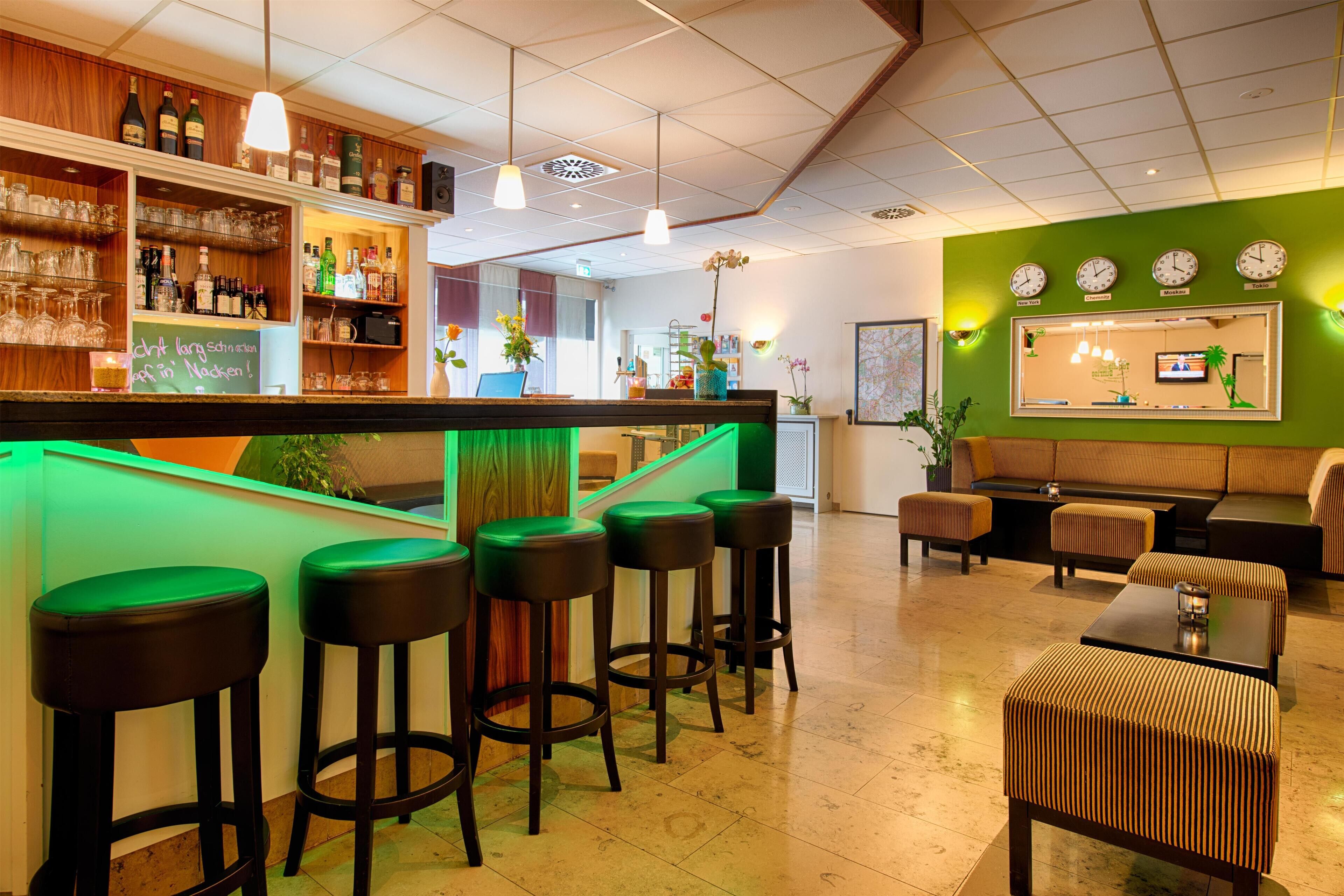Zona de bar con taburetes, mostrador y estanterías llenas de botellas y vasos. Hay una sala de estar con sofá y mesa.