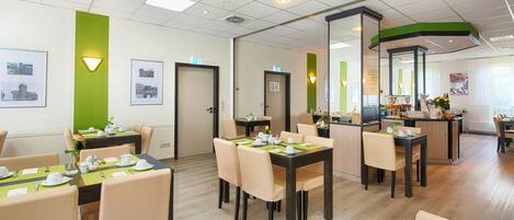 A hotel dining area with multiple tables and chairs, a kitchen area, and framed pictures on the walls.