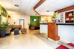 A hotel reception area with a green wall, wooden reception desk, seating area with sofas and chairs, and a television screen.