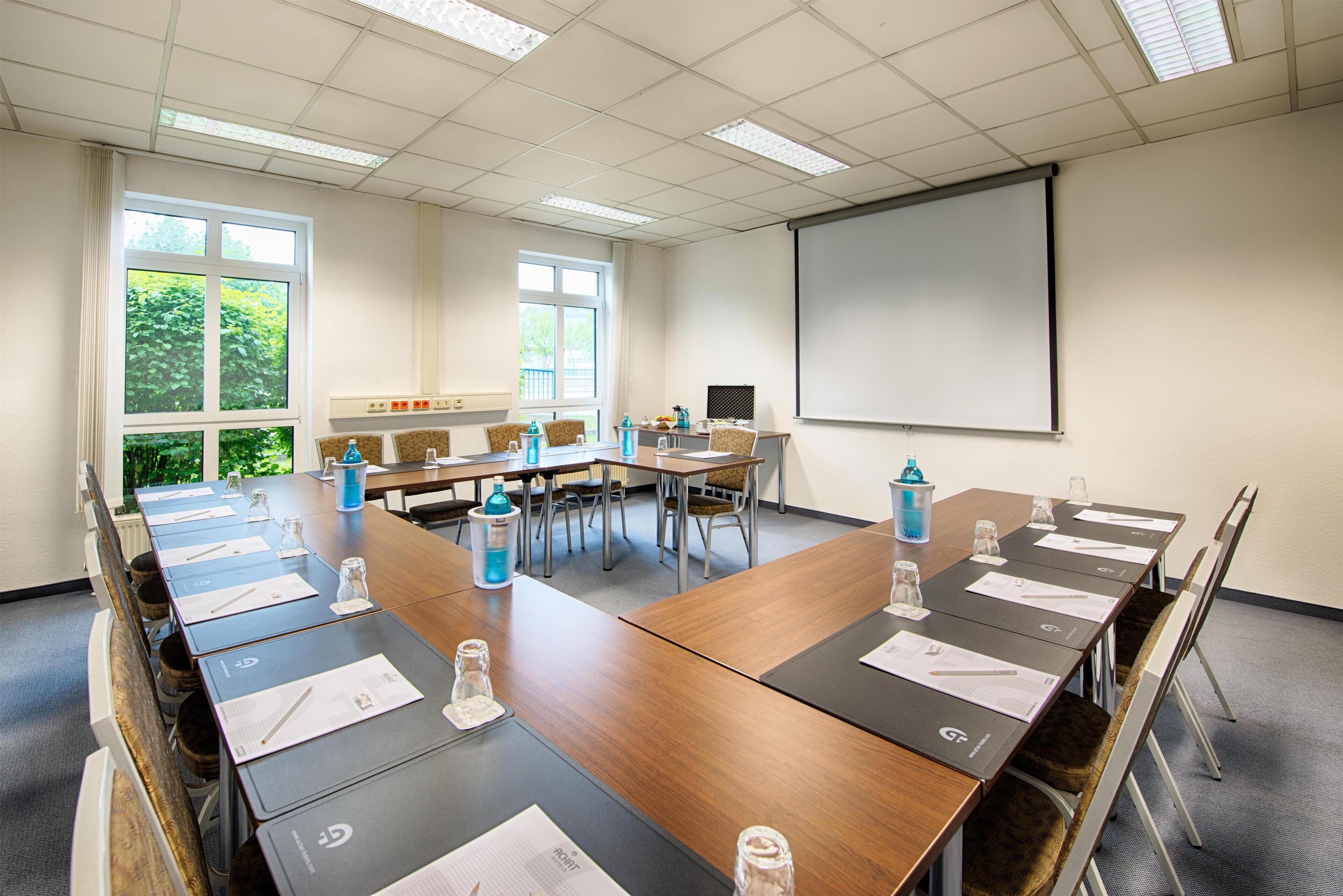 A conference room with a long table, chairs, a projector screen, and windows with a view of greenery.