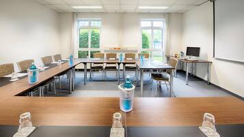 A conference room with a long table, chairs, a projector screen, and a window with a view of greenery.
