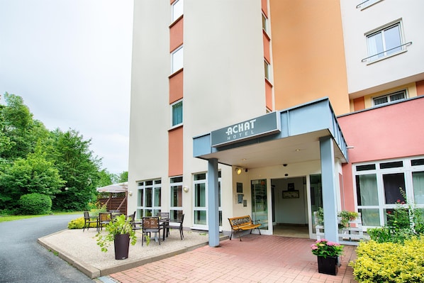 A hotel entrance with a sign reading "ACHAT HOTEL," outdoor seating, and potted plants.