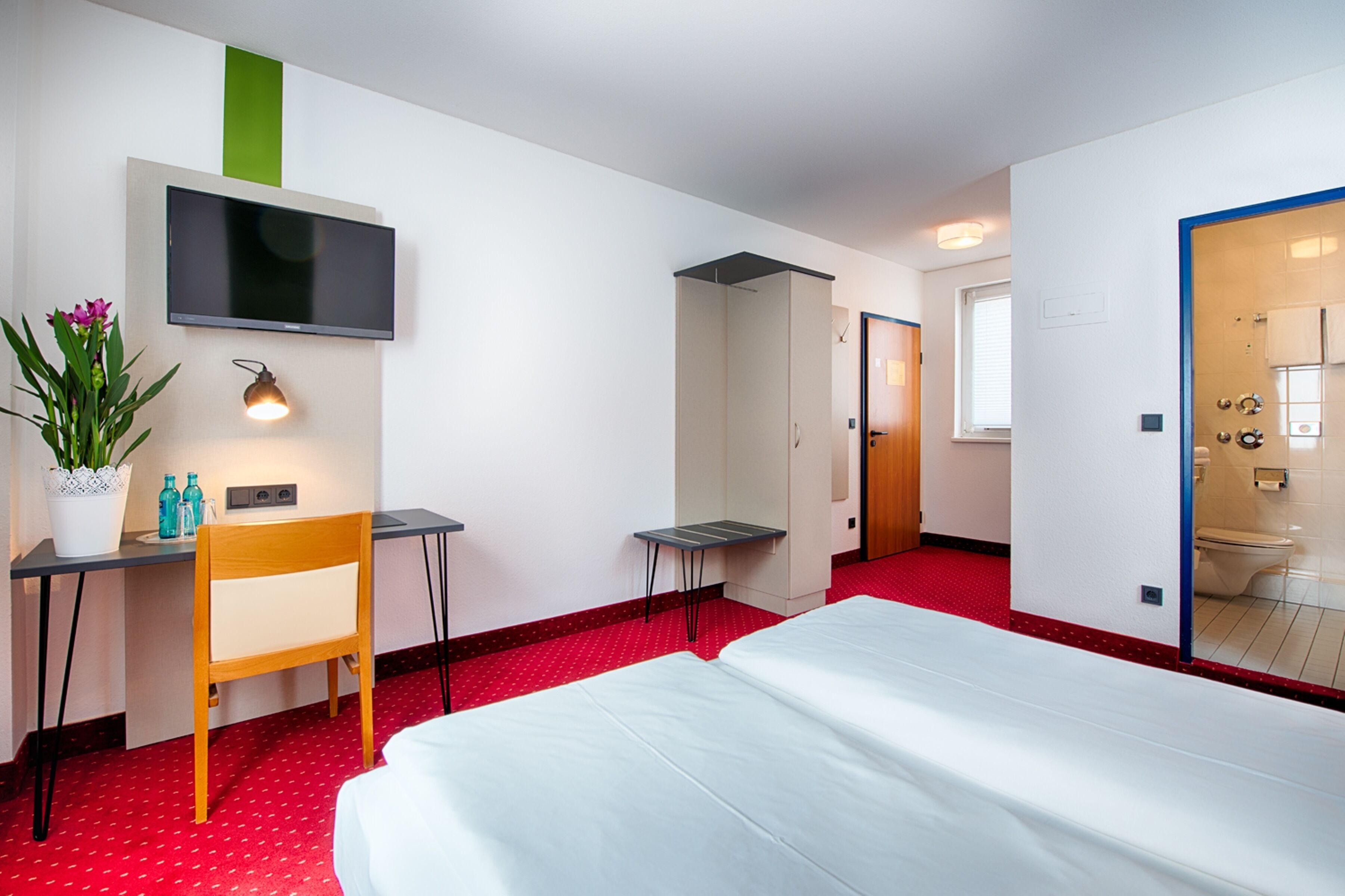 Quarto de hotel com cama, escrivaninha, cadeira, televisão e banheiro visível através de uma porta aberta.