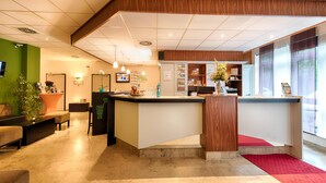 A reception area with a wooden reception desk, a seating area with a sofa and chairs, and a counter with a vase of flowers. - ACHAT Hotel Chemnitz (Chemnitz)