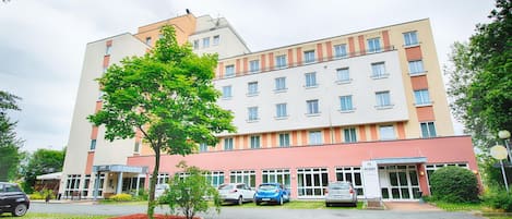 Un hôtel à plusieurs étages, doté d’un design moderne, avec une entrée principale, un parking pouvant accueillir plusieurs voitures et un jardin bien entretenu avec un arbre.