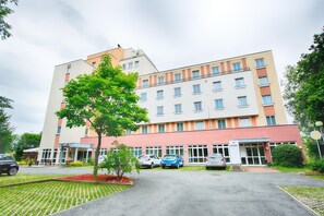 A multi-story hotel with a modern design, featuring a central entrance, a parking area with several cars, and a well-maintained garden with a tree.