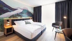 A hotel room with a bed, a desk, a chair, a nightstand, and a large mural on the wall.