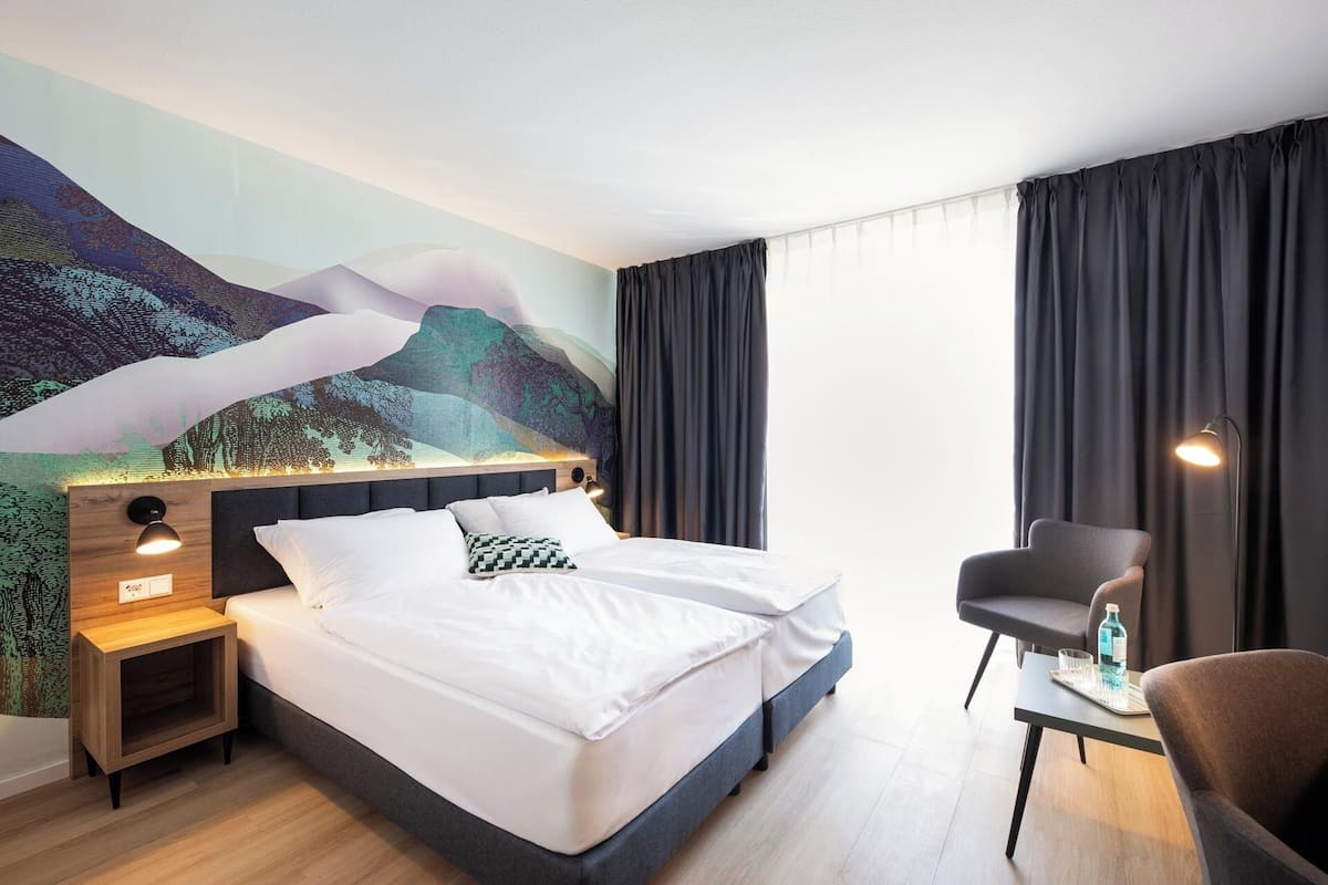 A hotel room with a bed, a desk, a chair, a nightstand, and a large mural on the wall.