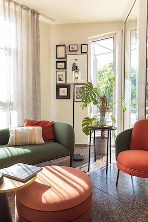 A modern living room with a green sofa, a red armchair, and a round ottoman. There is a floor lamp and a potted plant on a small table. - ACHAT Hotel Schwetzingen Heidelberg (Schwetzingen)
