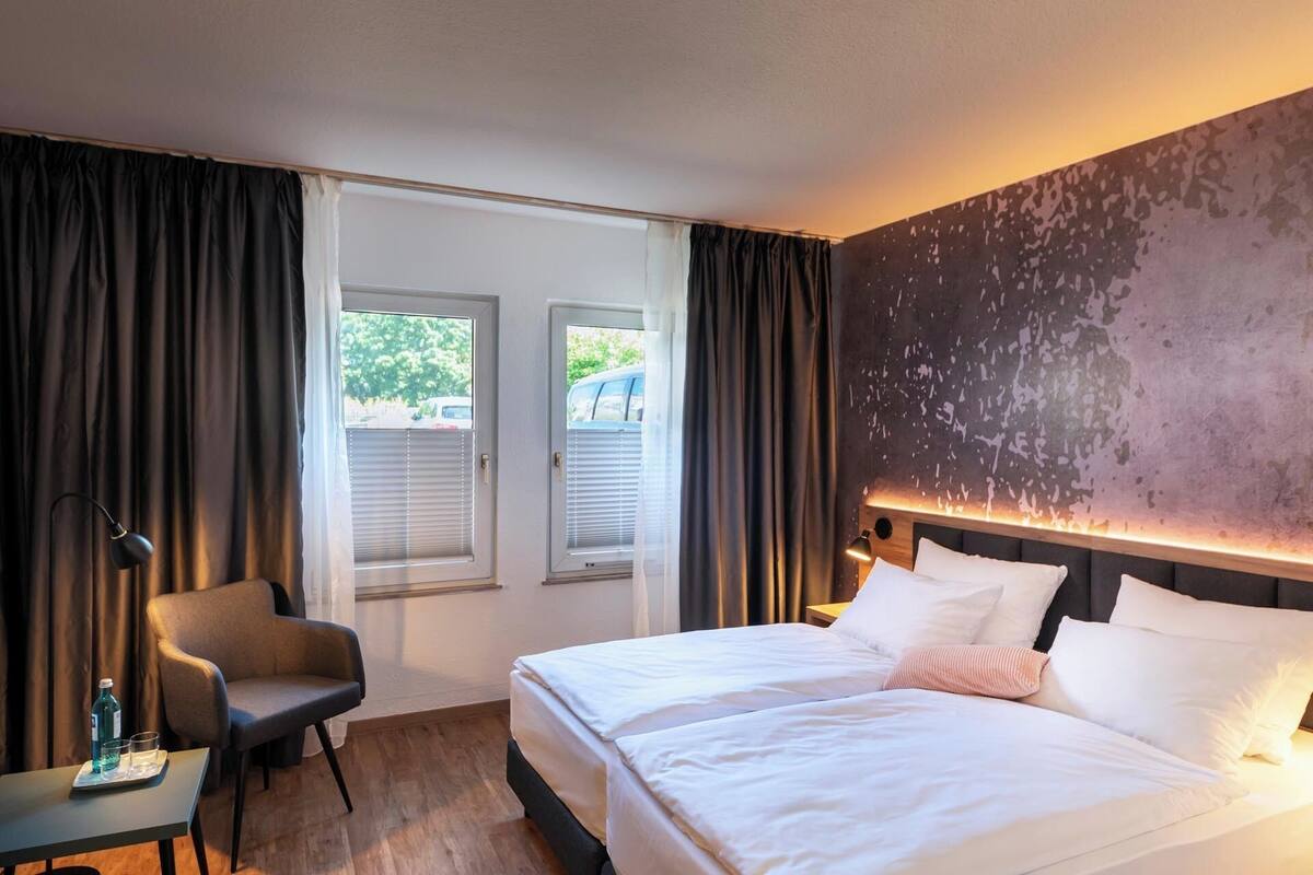 A hotel room with a bed, a chair, a table with a bottle and glasses, and a window with blinds.