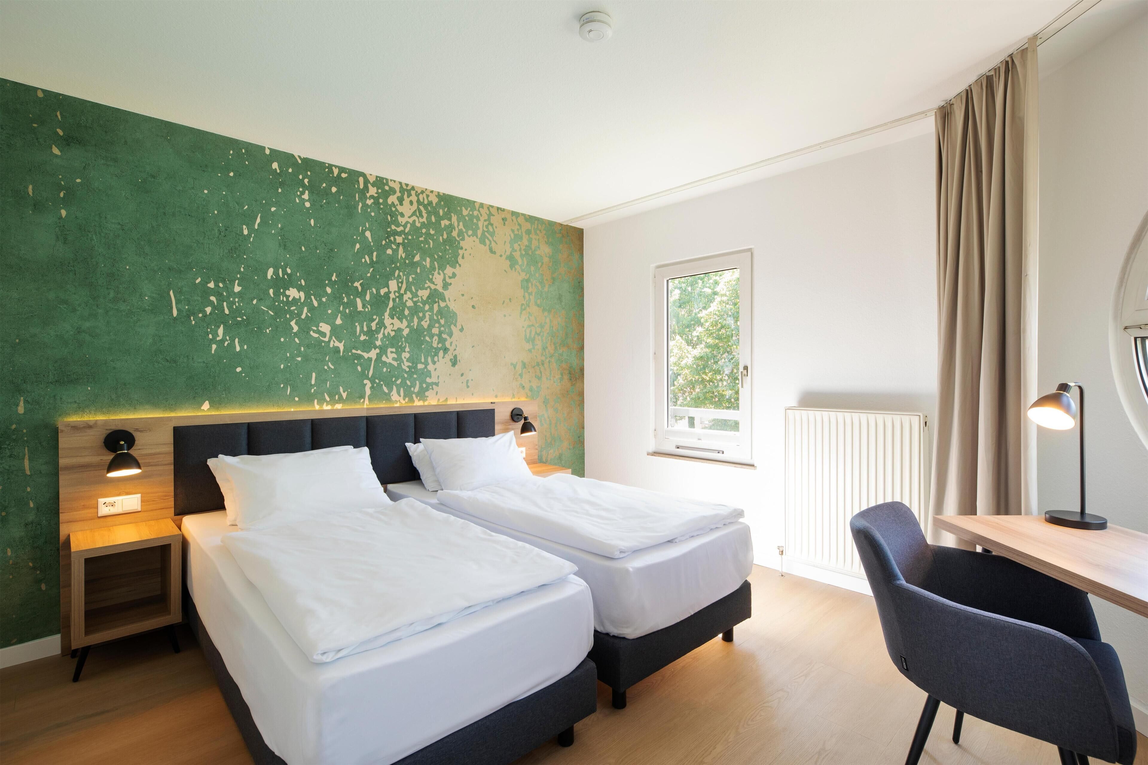 Une chambre d’hôtel avec deux lits, un bureau et une chaise. Il y a une fenêtre avec des rideaux, un radiateur et un mur dont la peinture s’écaille.