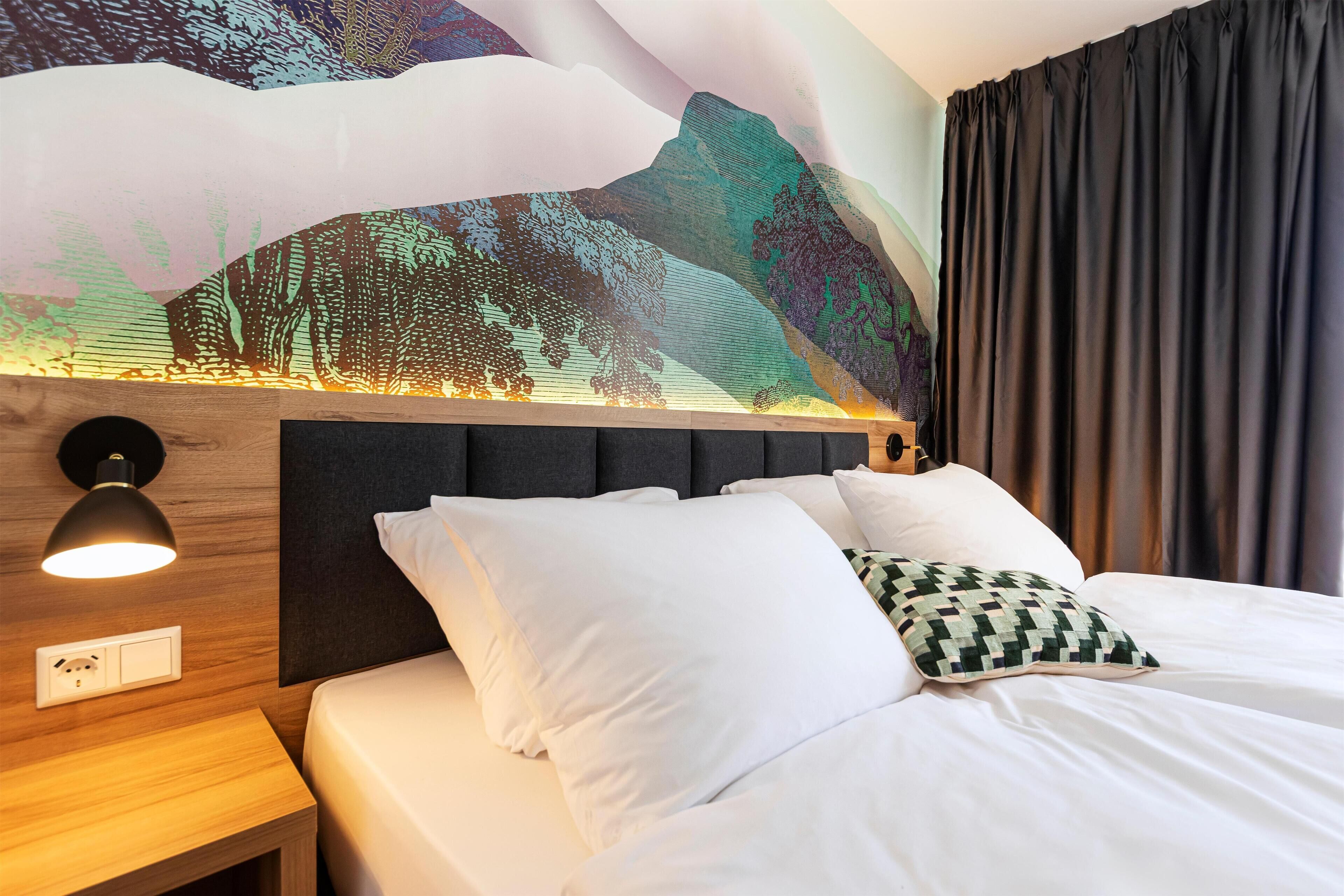 A hotel room with a bed, a wall-mounted lamp, a power outlet, and a large mural on the wall.
