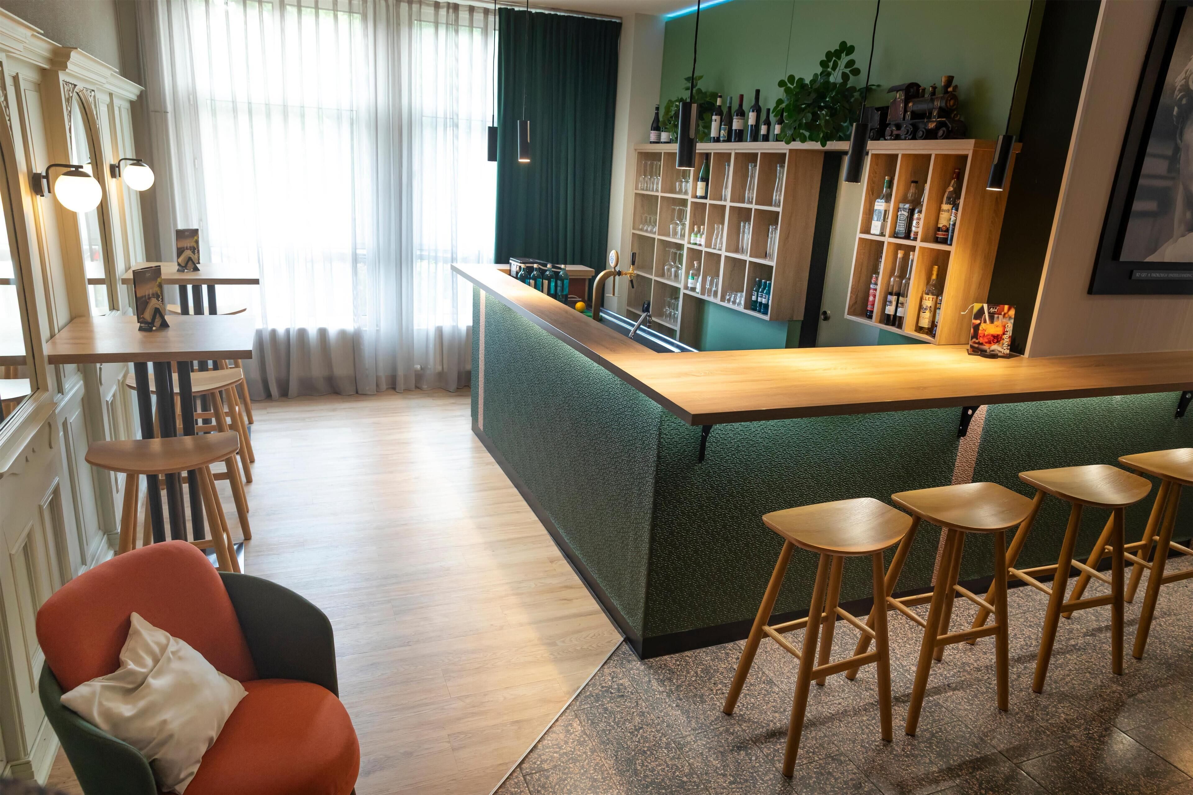 Un bar moderno con una barra acolchada de color verde, taburetes de madera y una estantería con botellas y vasos.