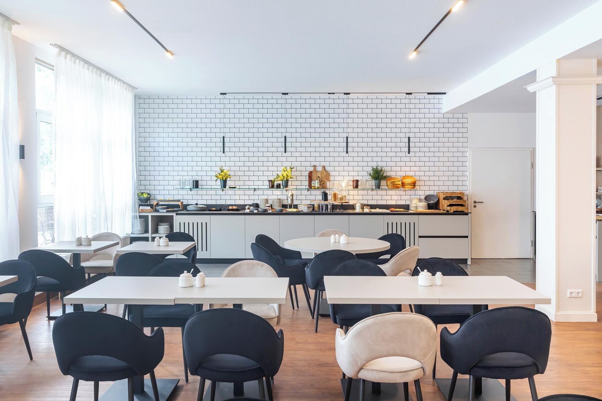 A modern dining area with white tables and chairs, a kitchen with white brick backsplash, and natural light from large windows.