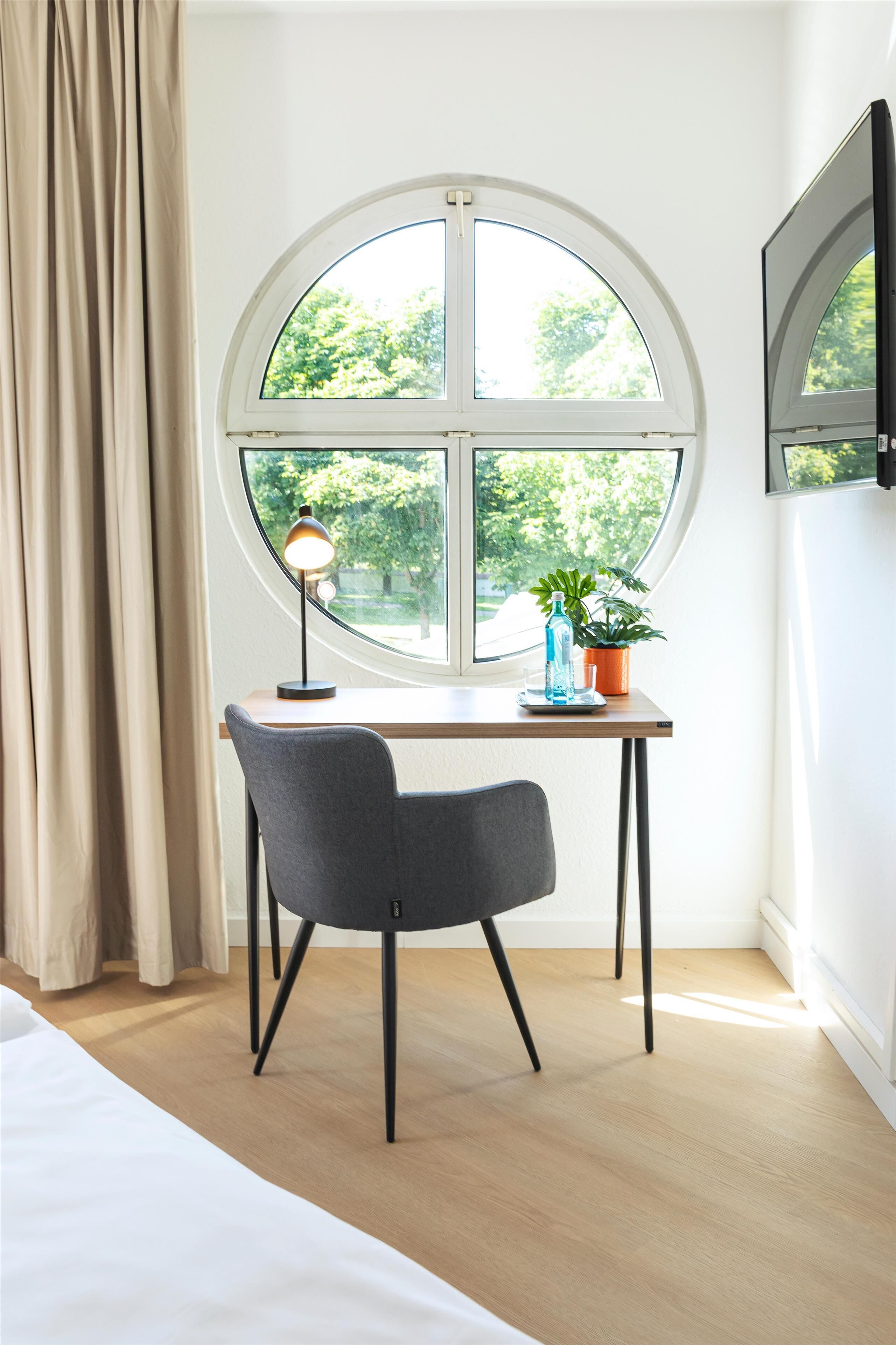 A room with a round window, a desk with a lamp, a chair, a potted plant, and a bed.