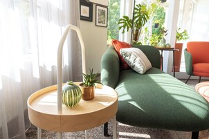 Un salón acogedor con un sofá verde, una mesita de madera con una planta, y una ventana con cortinas traslúcidas.