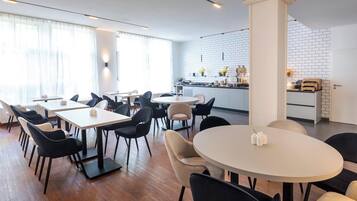 Un comedor moderno con mesas redondas y rectangulares, sillas negras y beige, y una cocina al fondo con paredes de ladrillo blanco y varios utensilios de cocina.