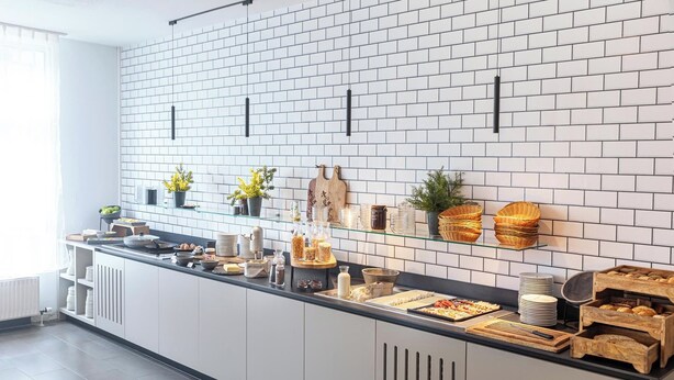 A modern kitchen with white subway tiles, a long counter with various food items, and a wooden cutting board.
