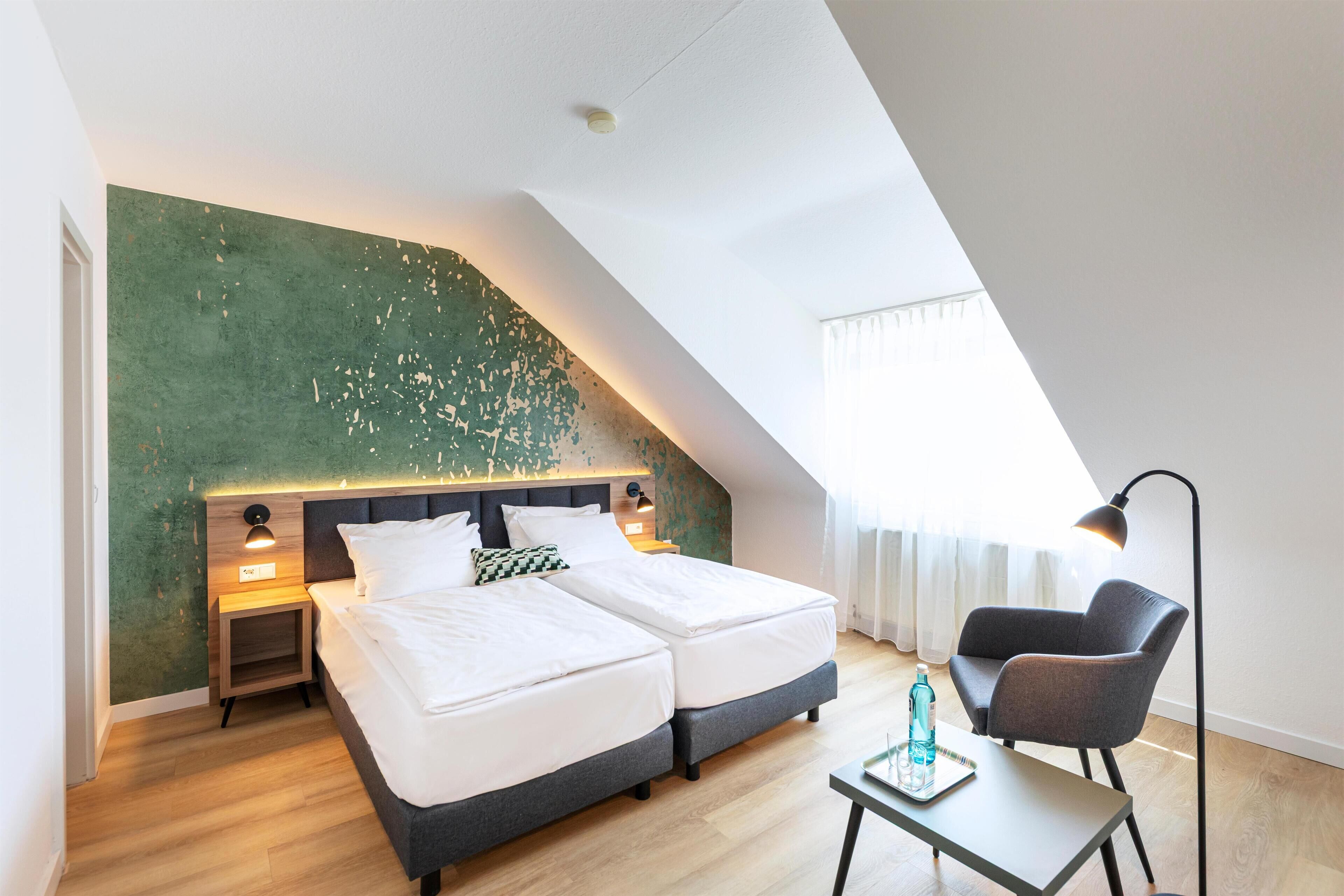 A modern hotel room with a sloped ceiling, two beds, a desk, a chair, and a large window.