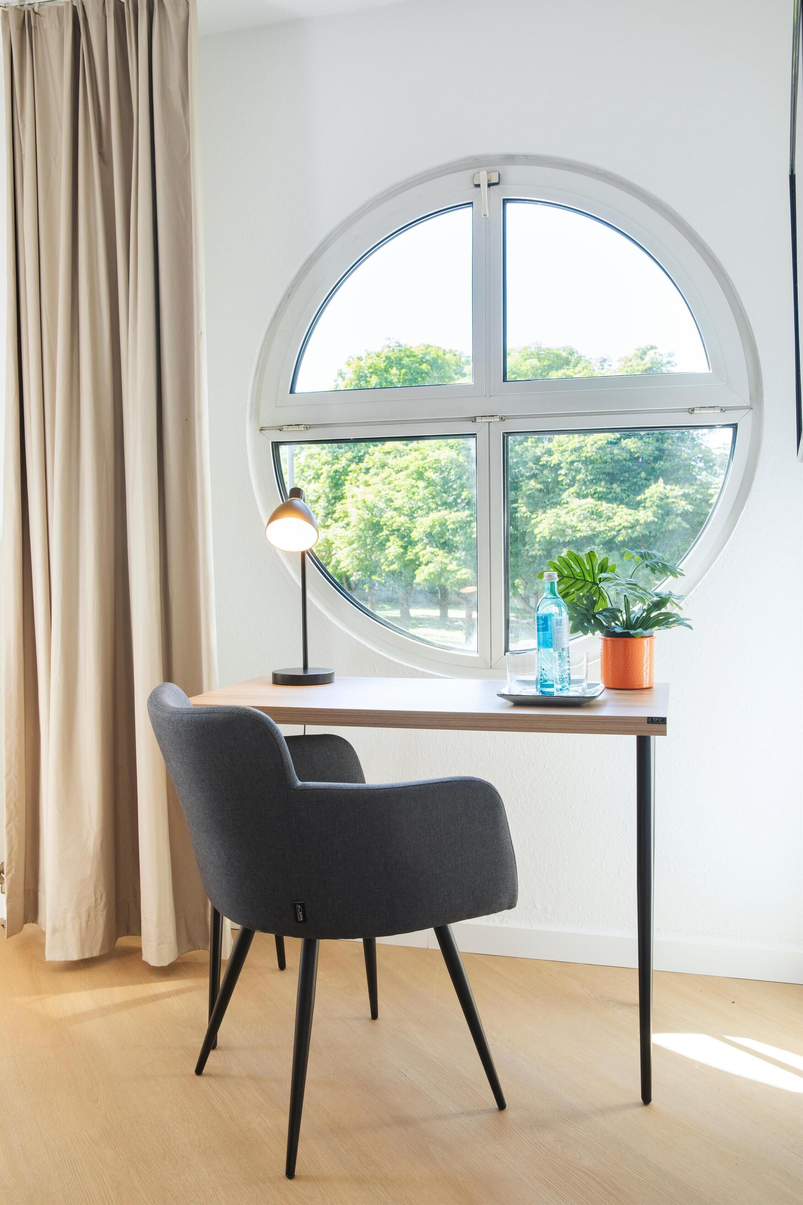 A room with a round window, a desk, a chair, a lamp, and a potted plant.