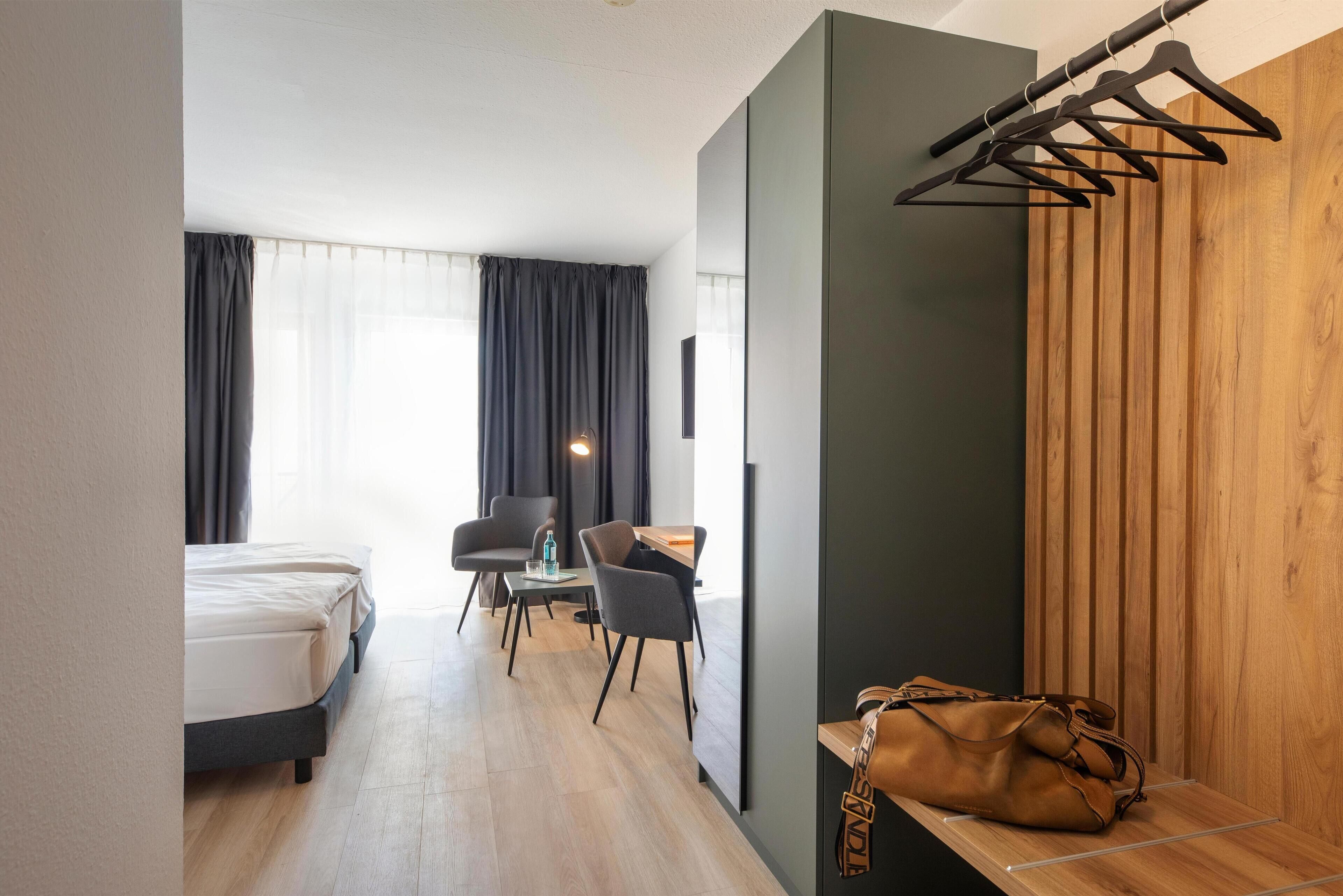 Ein Hotelzimmer mit einem Bett, zwei Stühlen, einem kleinen Tisch und einer Holzbank mit einer Tasche.