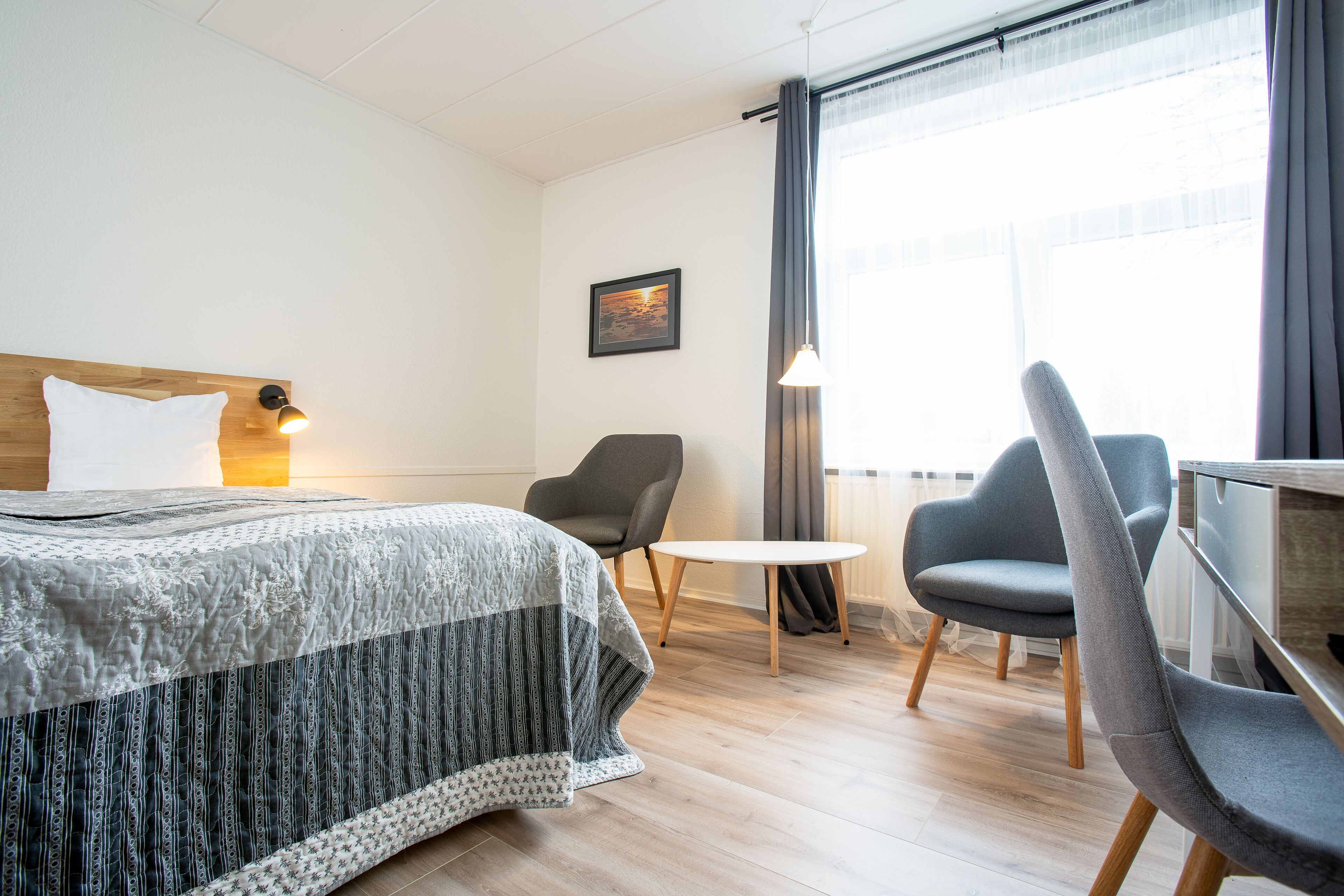 A hotel room with a bed, two chairs, a small table, a lamp, and a window with curtains.