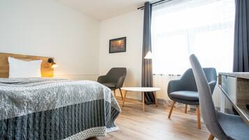 A hotel room with a bed, two chairs, a small table, a lamp, and a window with curtains.