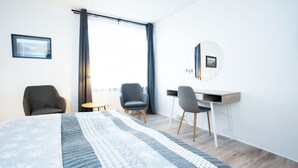 Une chambre d’hôtel moderne avec un grand lit, deux fauteuils, un bureau et une grande fenêtre avec des rideaux.