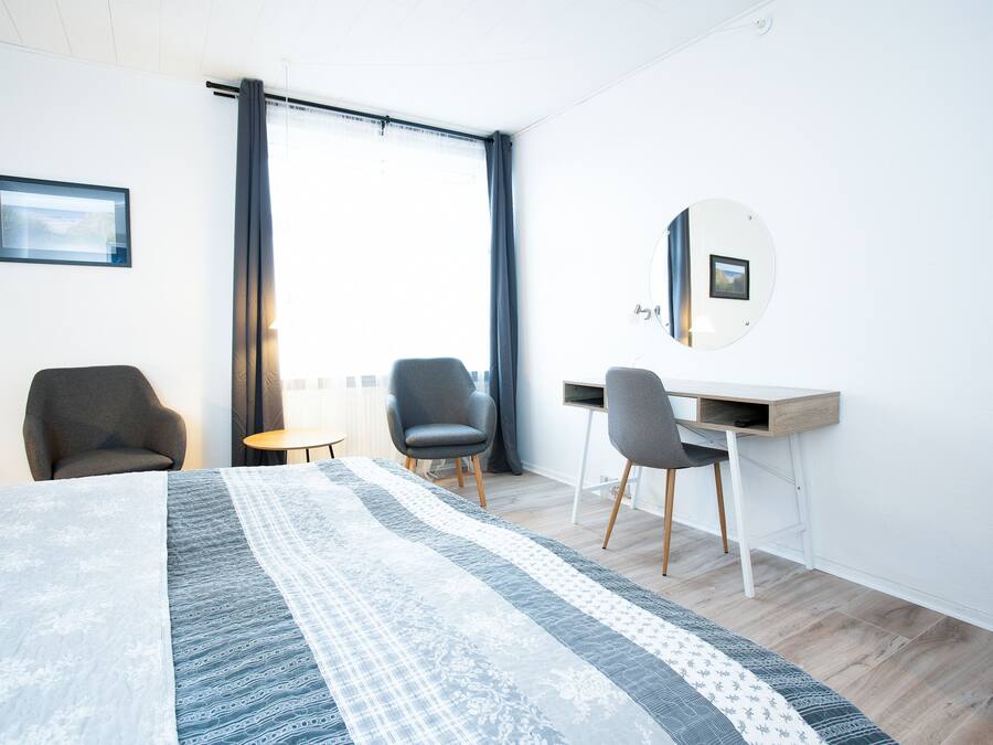 Una habitación de hotel moderna con una cama grande, dos sillas, un escritorio y un ventanal con cortinas.