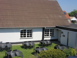A building with a corrugated metal roof, white exterior walls, and a small attached structure with a white exterior. There are outdoor tables and chairs arranged on a grassy area in front of the building.