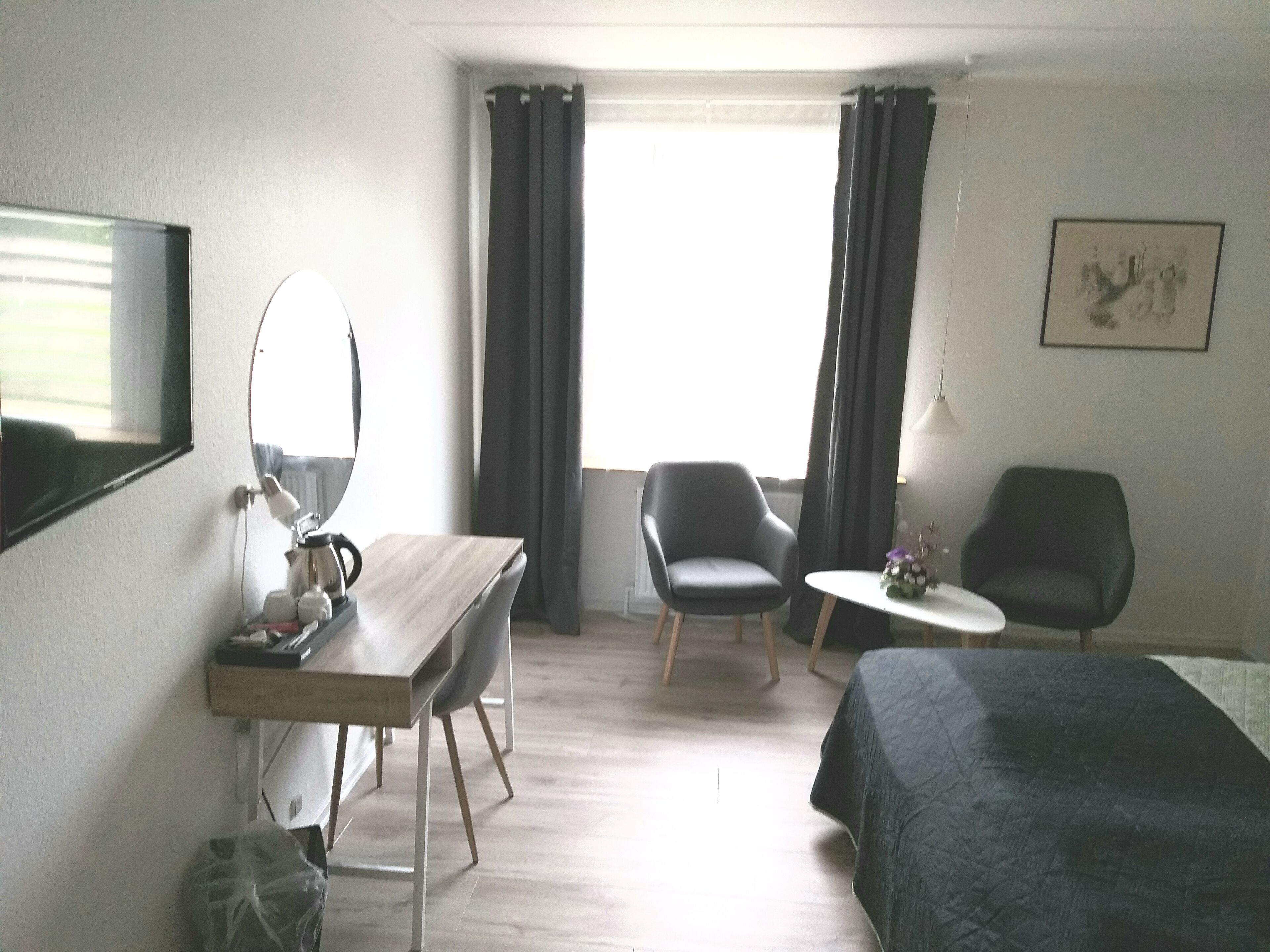 Habitación de hotel con cama, dos sillas, un escritorio con espejo, una mesita de noche con lámpara y una ventana con cortinas.