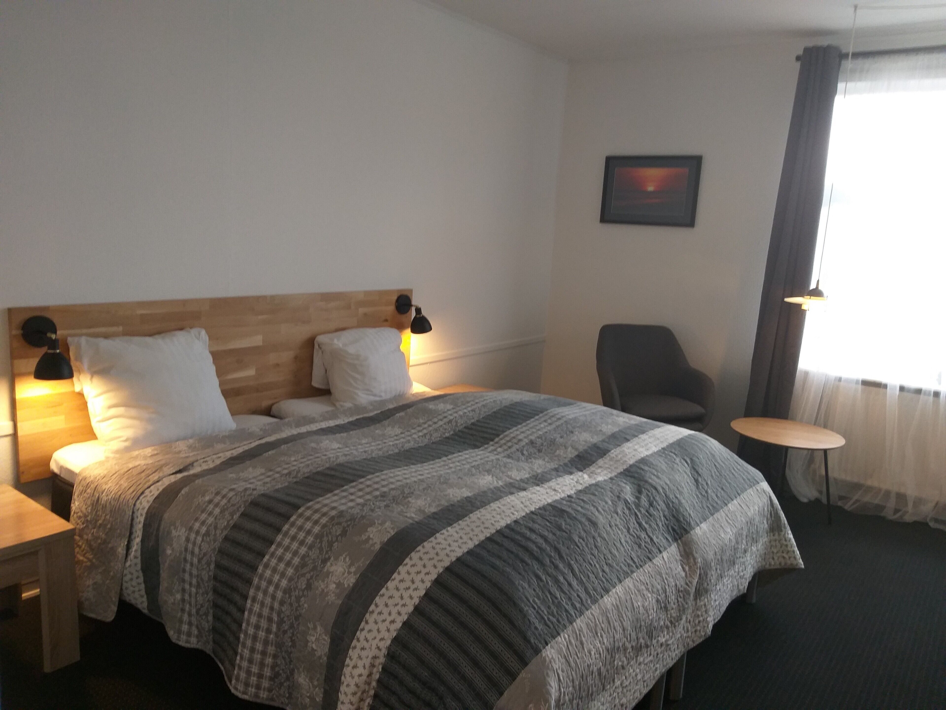 Una cama bien hecha con una manta a rayas, un cabecero de madera, dos lámparas de noche, una silla, una mesita y un cuadro en la pared.